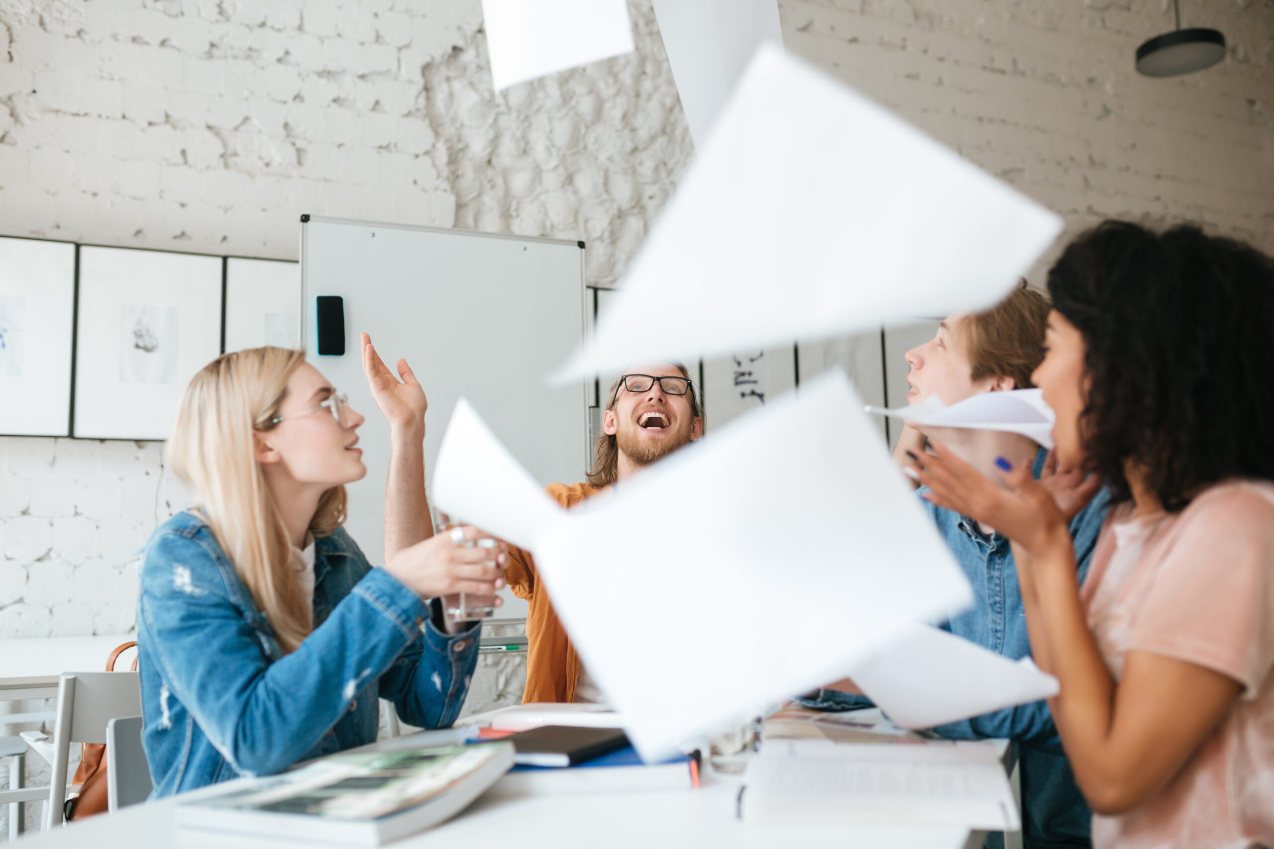emotional people happily throw up papers in office while working together. group of young students joyfully studying with books on table in classroom.